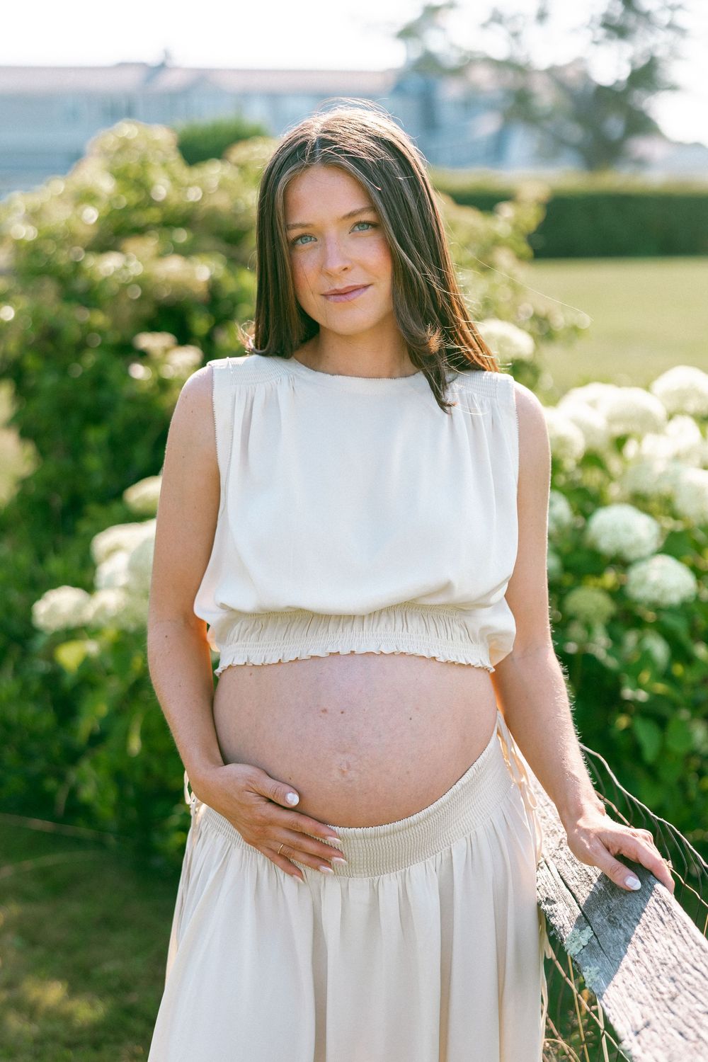 Pregnant woman in white crop top and skirt cradling her baby bump outdoors among white hydrangeas.