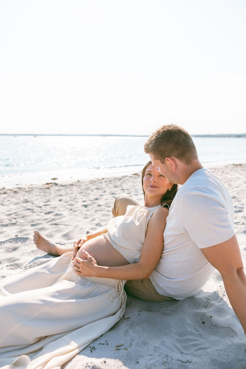 Pregnant couple sitting together on sandy beach, man embracing woman from behind near ocean shoreline.