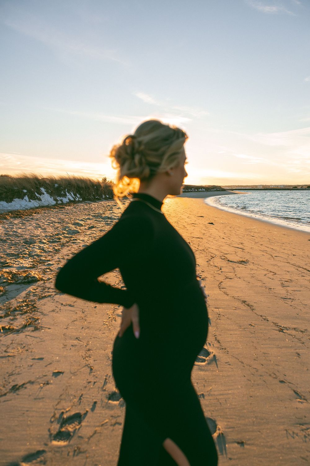 Silhouette of pregnant figure in black dress standing on beach at sunset with golden light illuminating the scene.