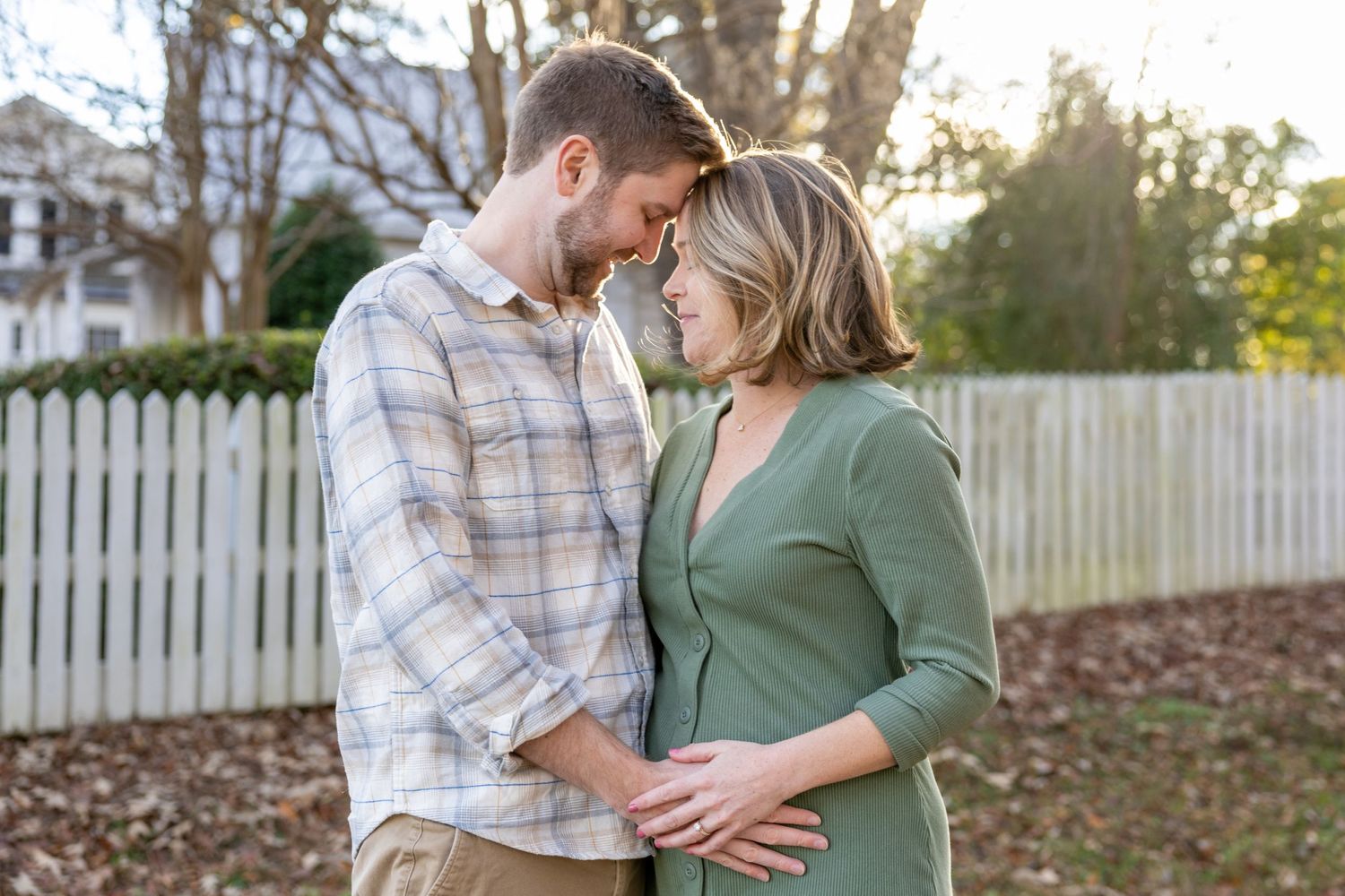 A couple stands affectionately in front of a white picket fence during a golden sunset in their backyard.