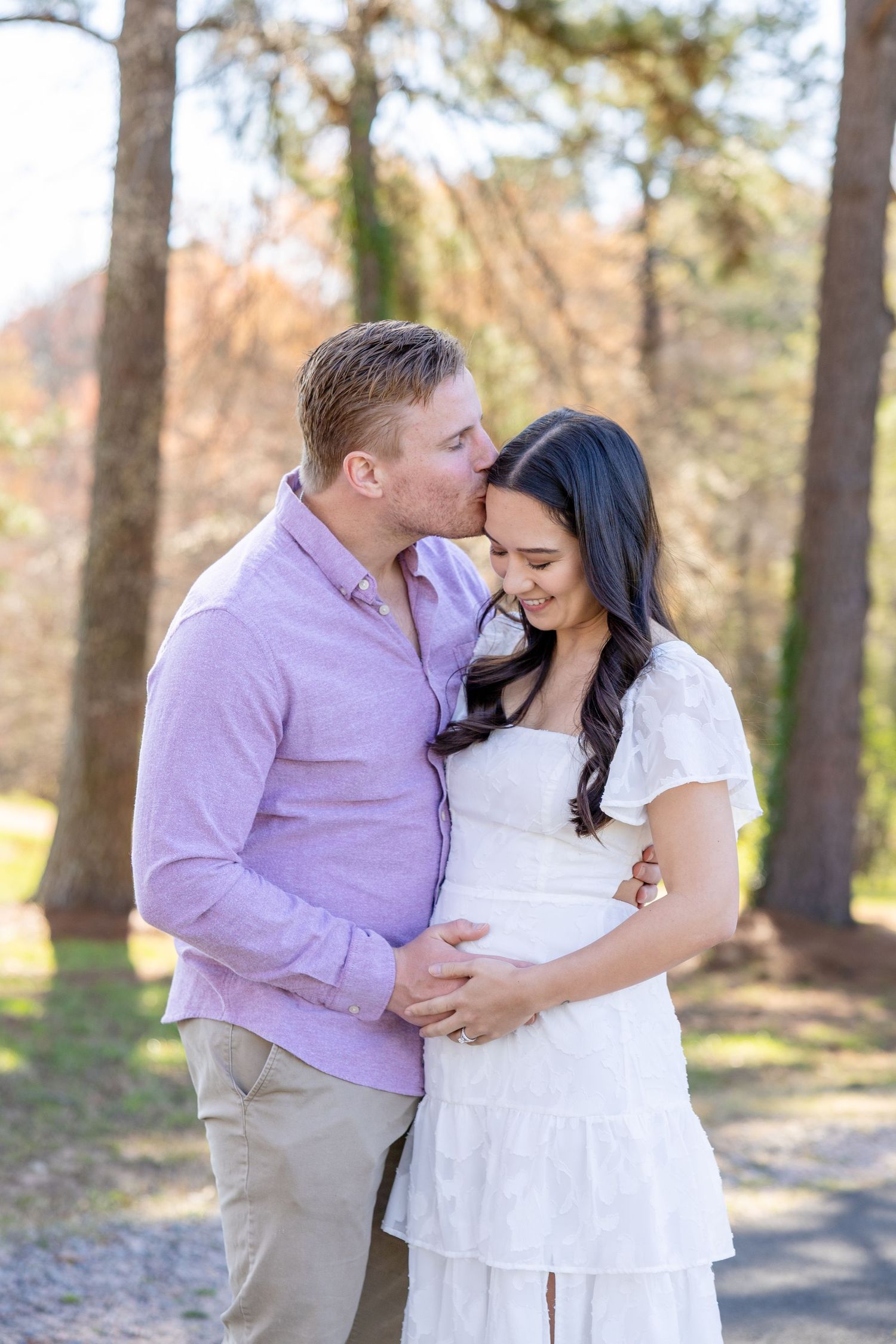 Couple standing in sunny forest setting during maternity photo shoot wearing pink and white outfits at the NC Art Museum in Raleigh.
