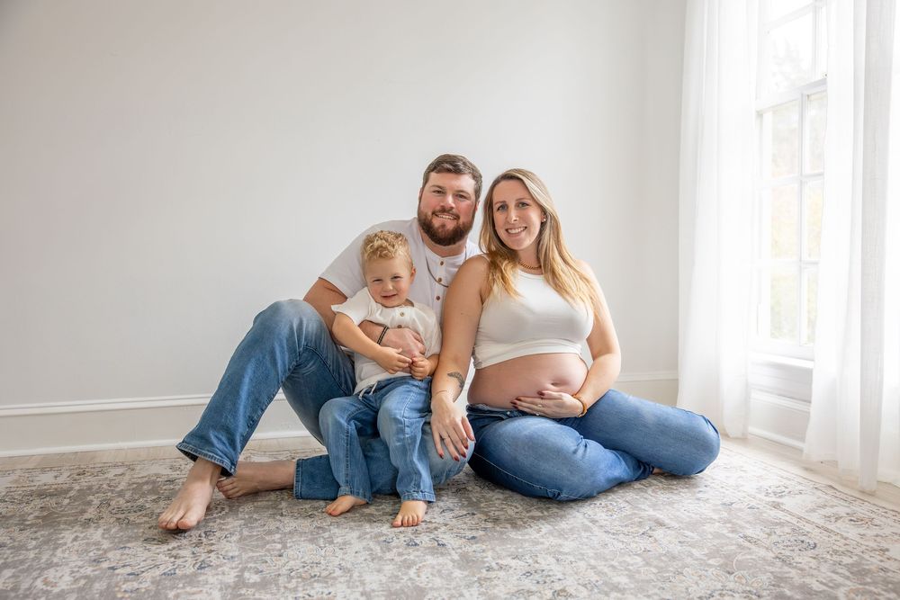 Pregnant woman sits with husband and toddler on carpet floor against white wall for maternity family photo.