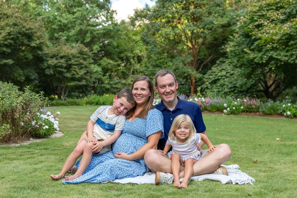 Happy family of four sitting on grass in a lush garden, pregnant mother smiling with husband and two young children.