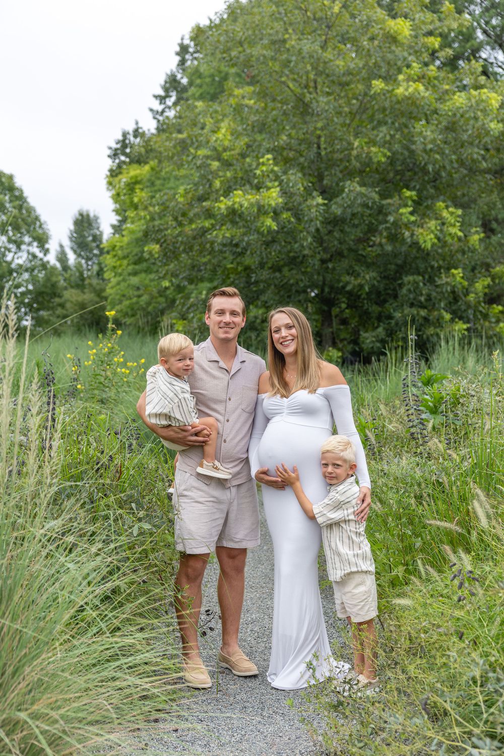 Pregnant woman in white dress with husband and two young boys on a garden path surrounded by lush greenery - Raleigh Maternity session