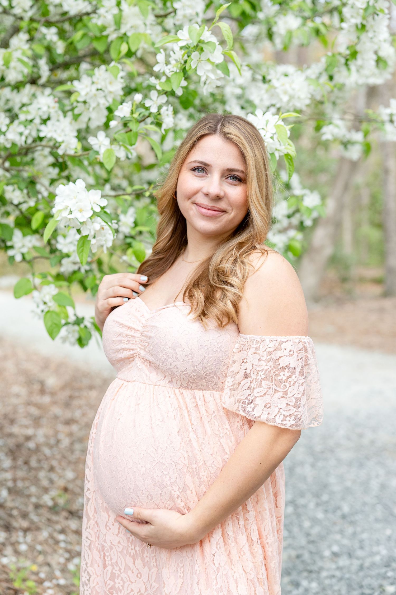 A pregnant woman in a pale pink lace dress poses by blooming white spring flowers on a gravel path.