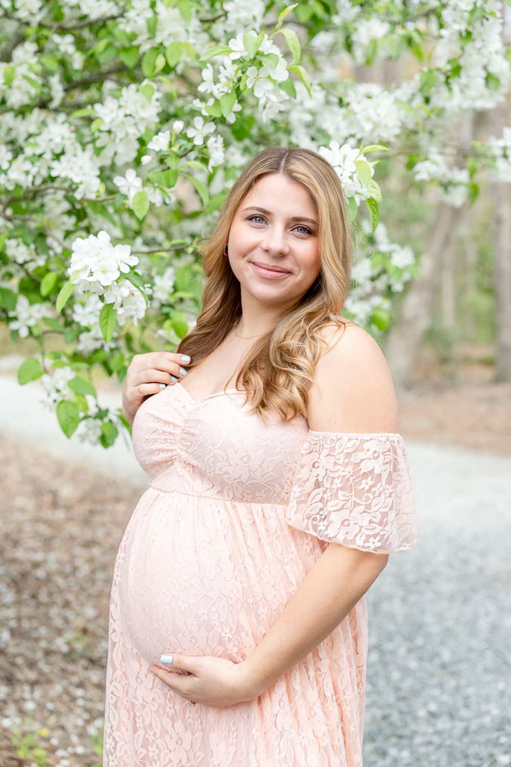 A pregnant woman in a pale pink lace dress poses by blooming white spring flowers on a gravel path.