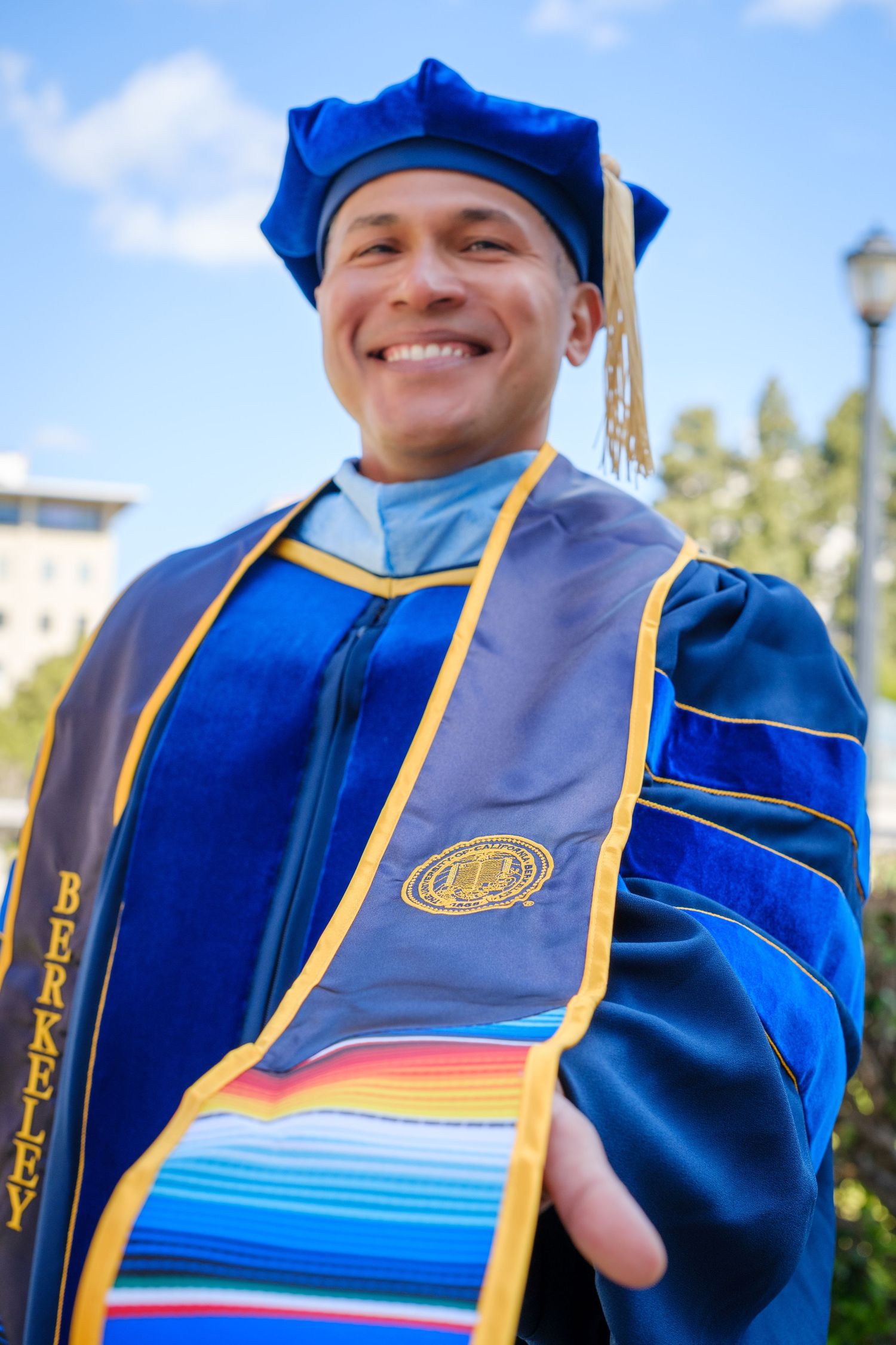 Smiling graduate in blue doctoral regalia and Berkeley stole poses outdoors at UC Berkeley commencement.