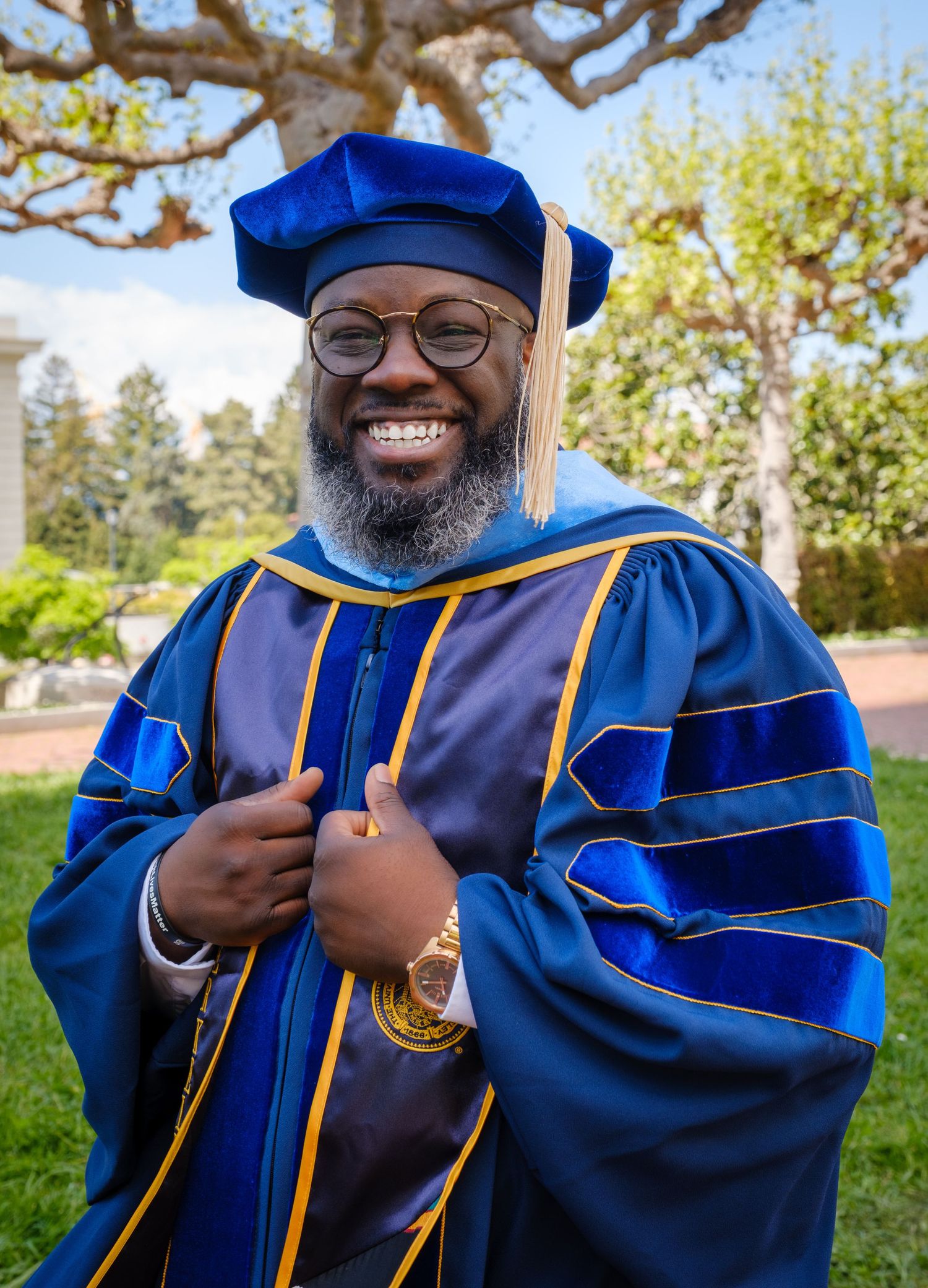 Smiling graduate in blue and gold doctoral regalia and cap poses outdoors on a sunny campus.