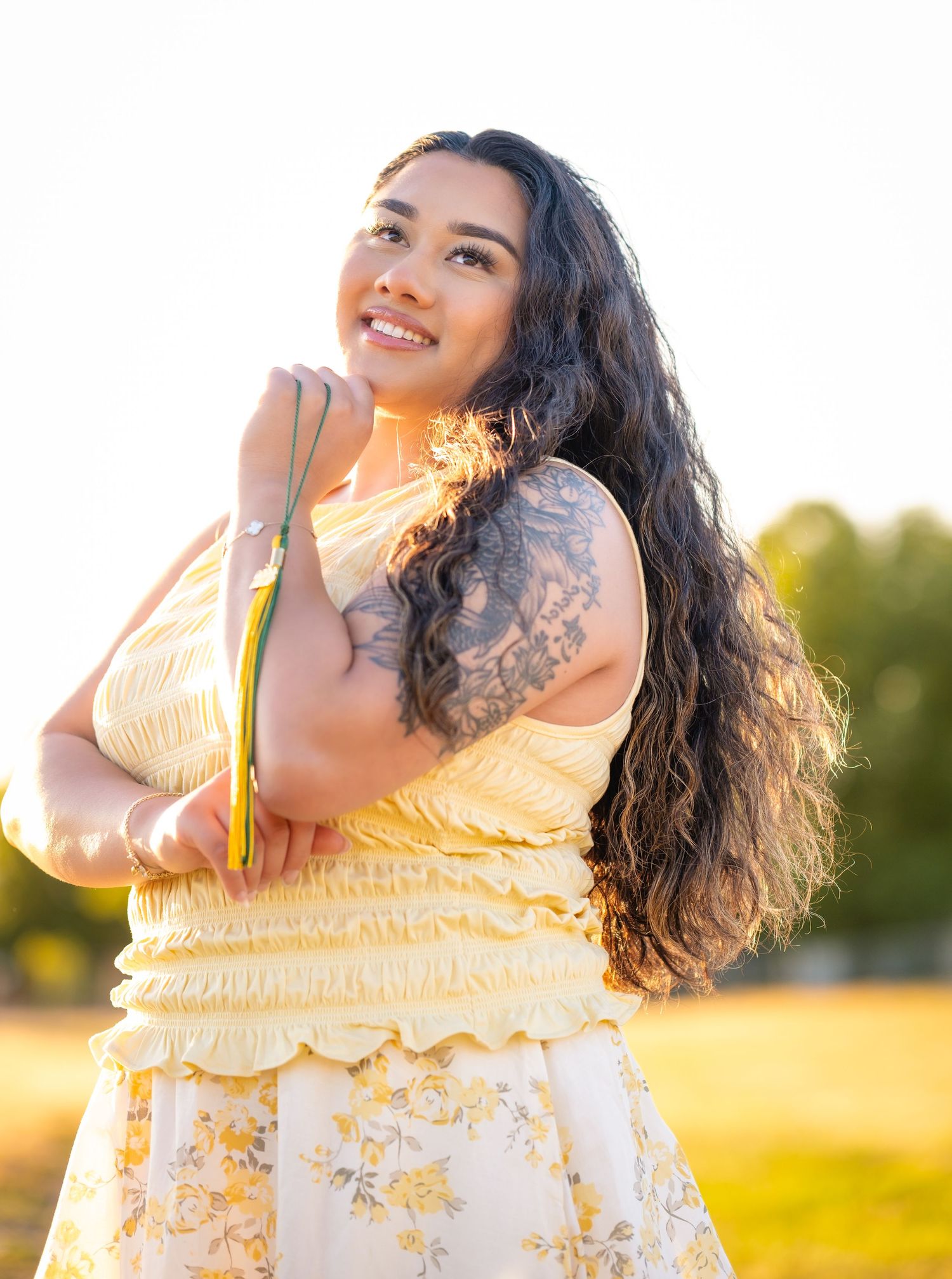 Young woman in yellow floral dress with tattoo smiling outdoors in golden sunlight.