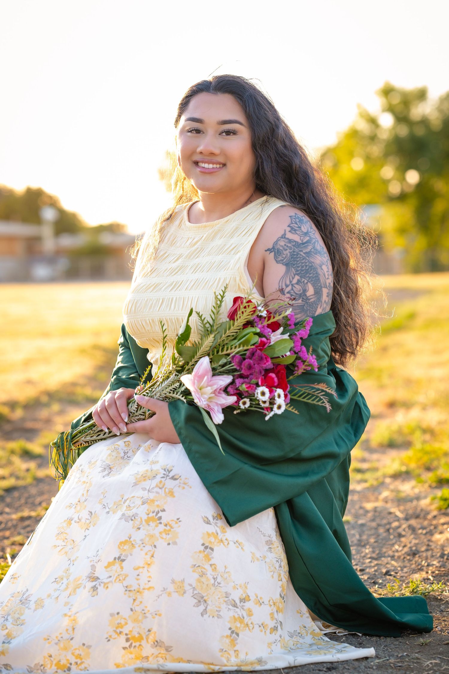 Young woman in floral gown and green sash holds colorful bouquet, smiling outdoors at golden hour.