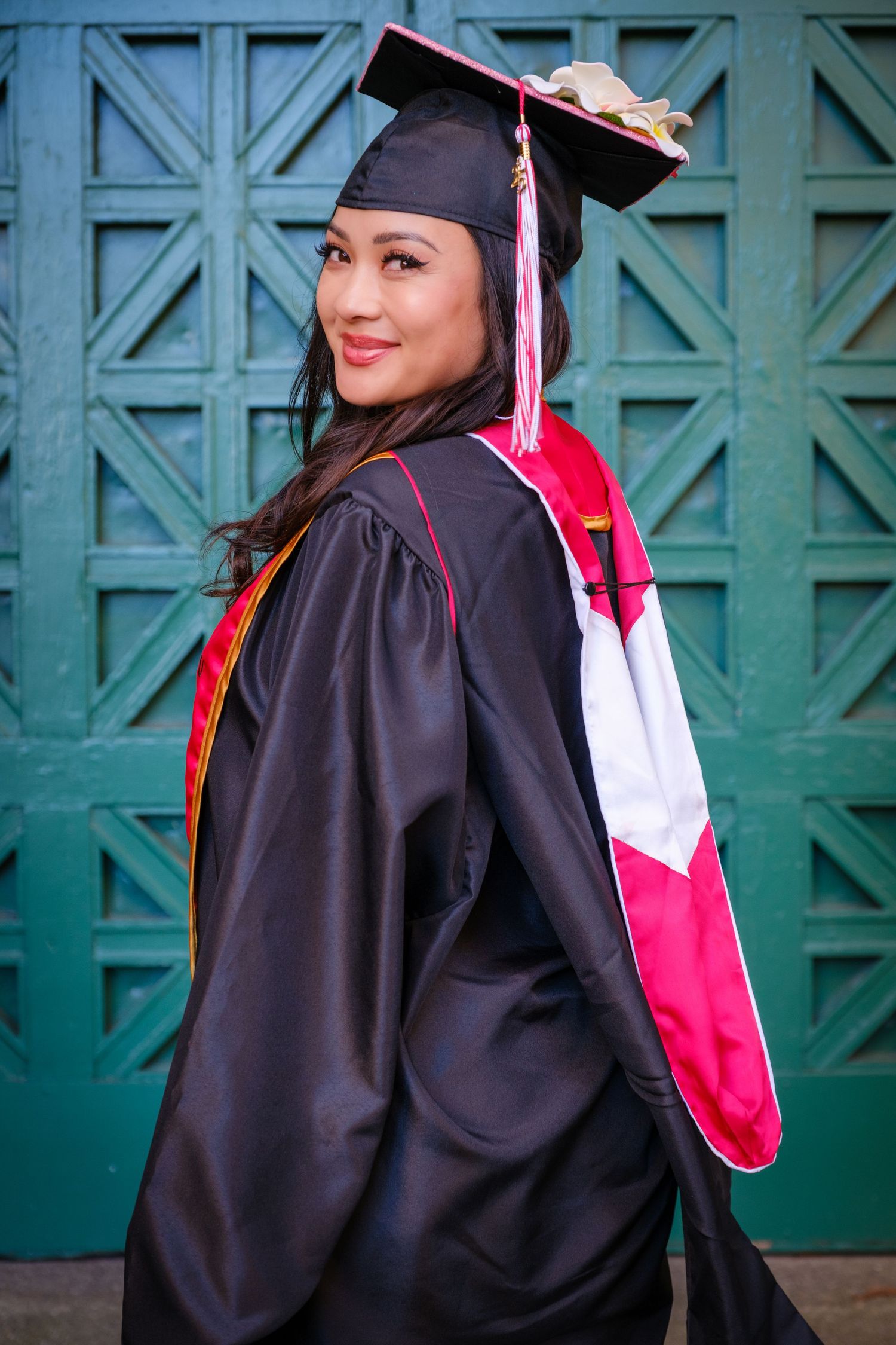 Smiling graduate in black cap and gown with pink and red sash posing outdoors near teal lattice wall.