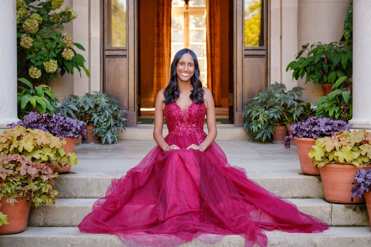 Young woman in a stunning fuchsia ball gown seated on stone steps surrounded by lush greenery and purple flowers.