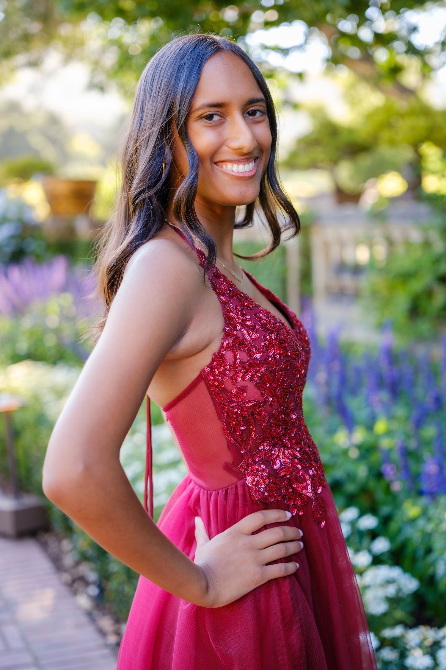 A smiling young woman in a red sequined halter gown poses outdoors in a garden setting.