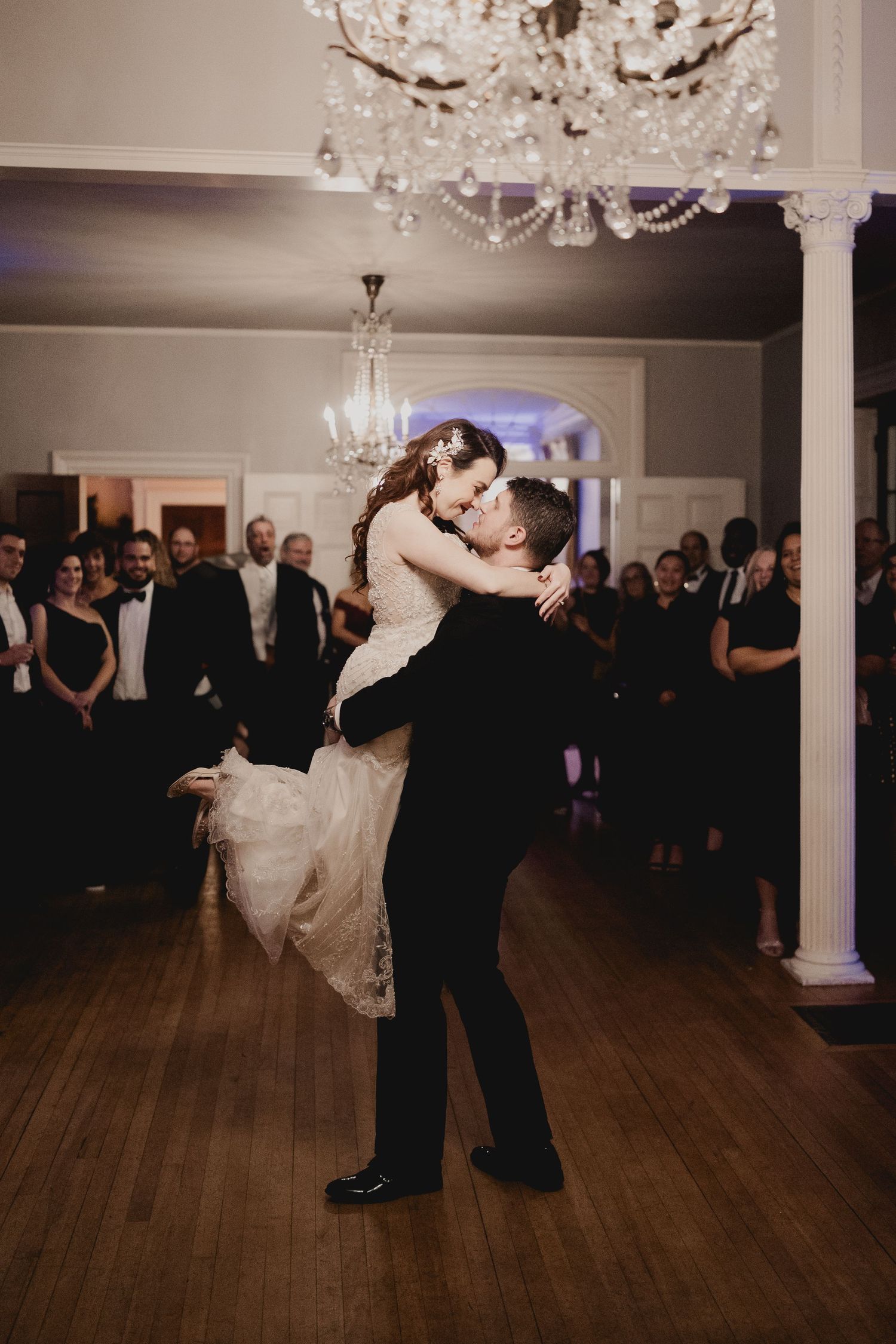 A couple shares their first dance under crystal chandeliers in an elegant ballroom with wooden floors.