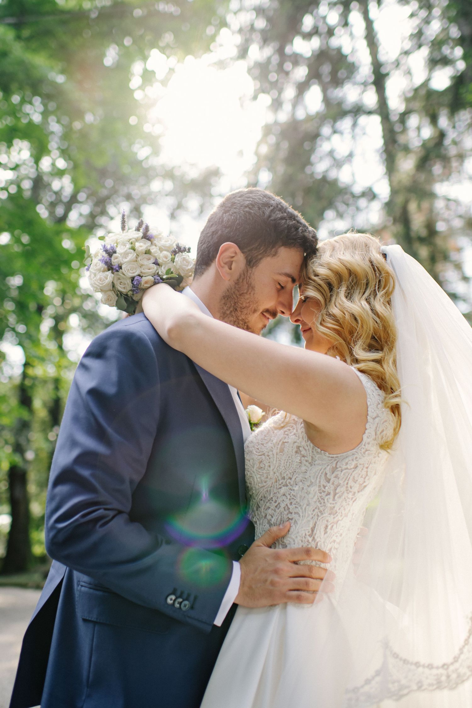 A romantic wedding couple embraces in a sun-dappled forest setting with soft natural lighting and flowing white veil.
