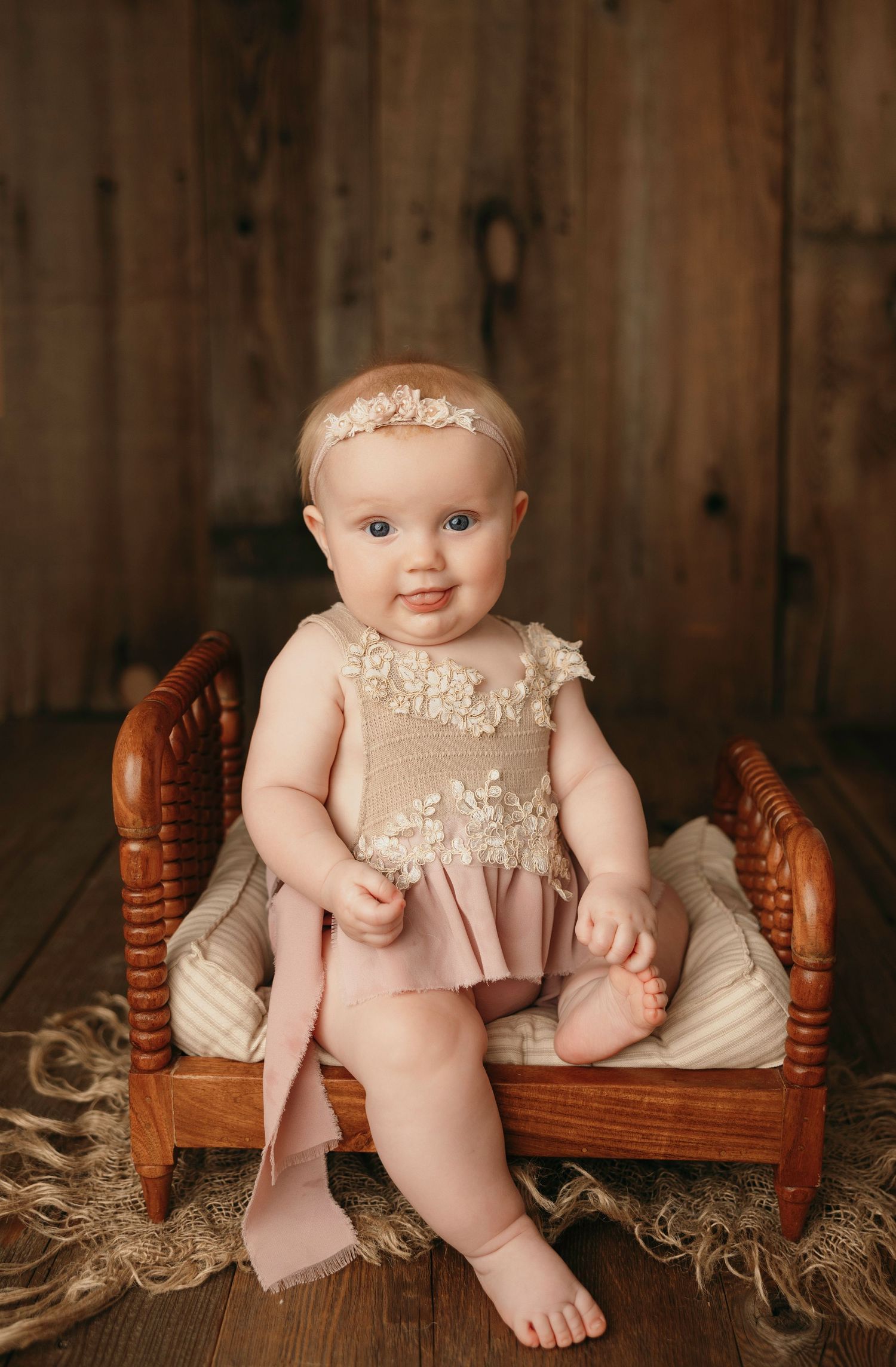 Baby wearing delicate lace dress sits on small wooden canopy bed with white linens against rustic wood backdrop.