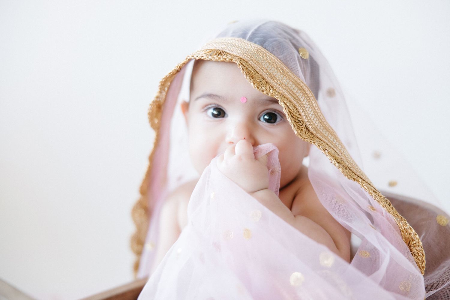 A baby wearing a woven straw hat peeks through soft pink fabric with a gentle expression.