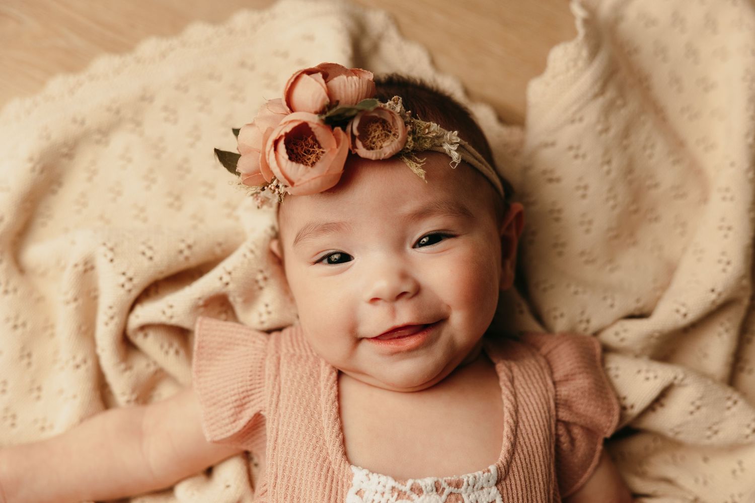 Baby wearing pink floral headband and ruffled outfit lies on cream colored blanket while smiling.