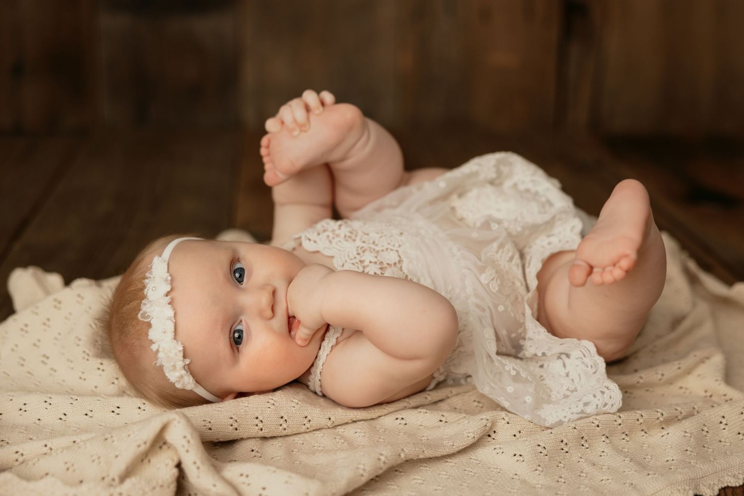 Infant wearing delicate white lace dress and headband lying on beige blanket during professional photo session.
