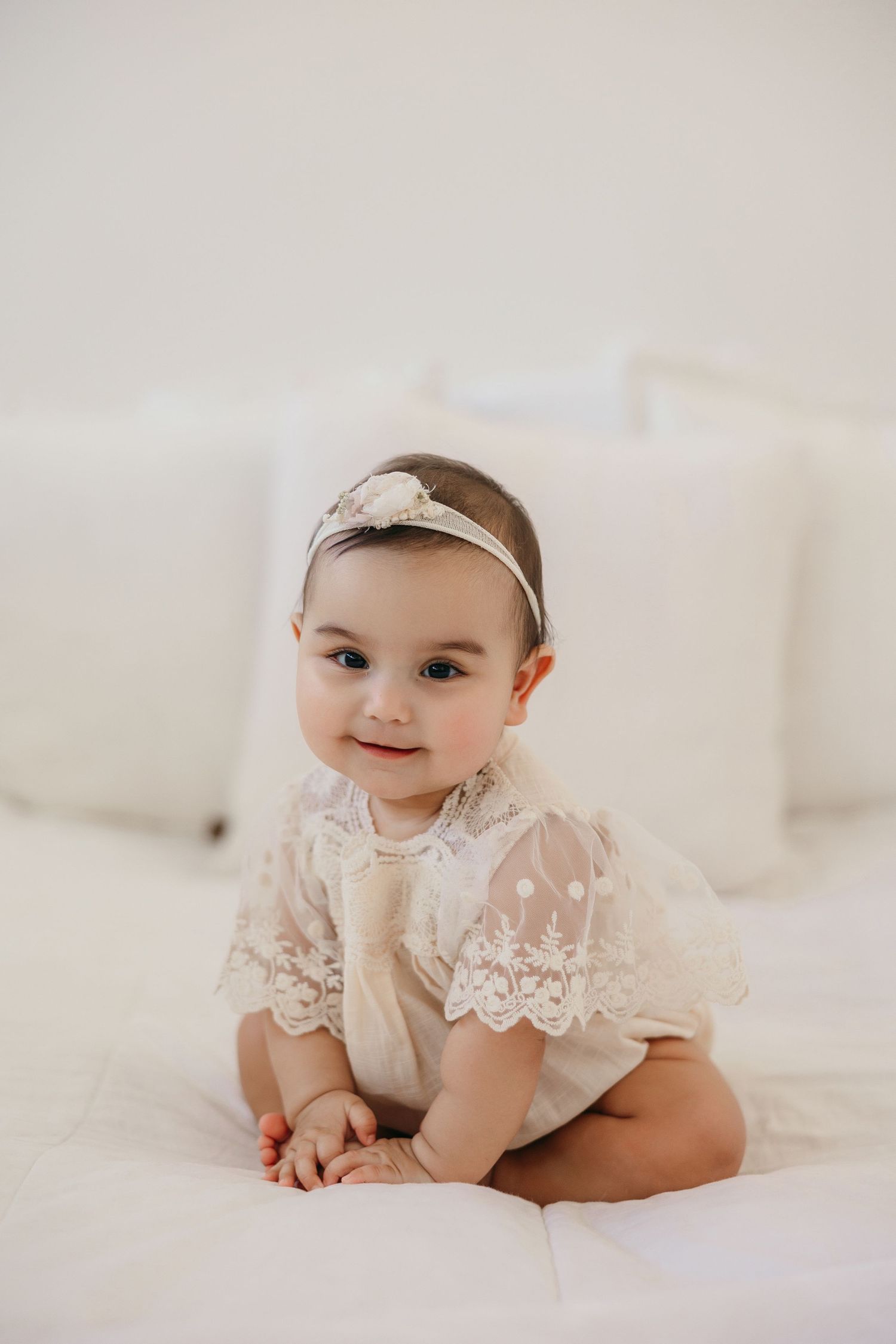 Baby wearing delicate white lace dress and headband sits on white bedding in soft neutral portrait setting.