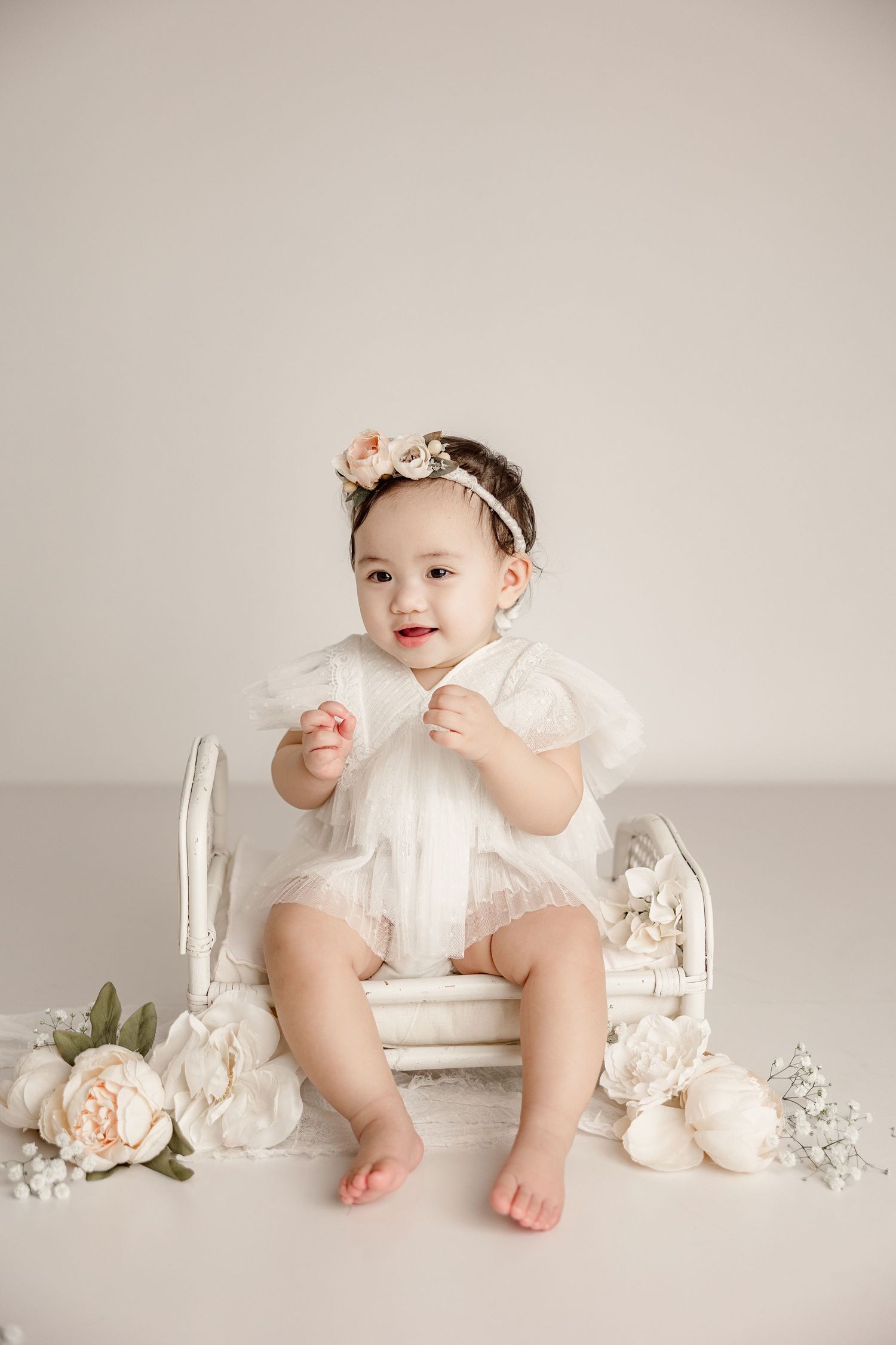 Baby wearing white dress sits on vintage chair with floral decorations in soft neutral studio setting.