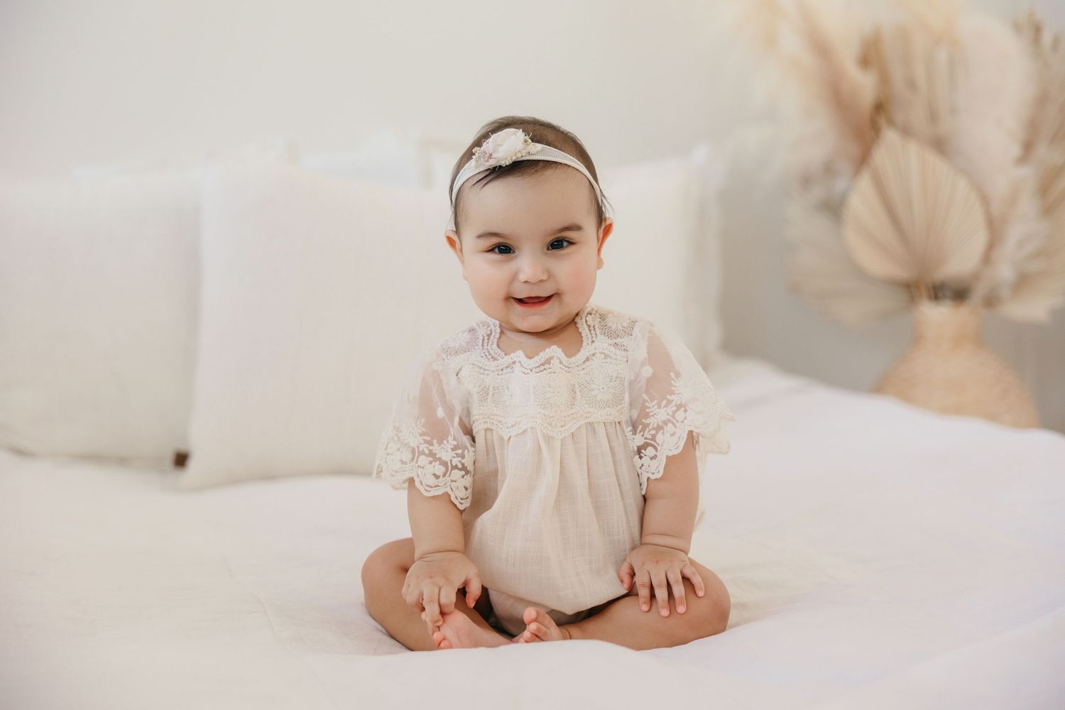Baby wearing delicate white lace dress sits on white couch with warm neutral decor in background.