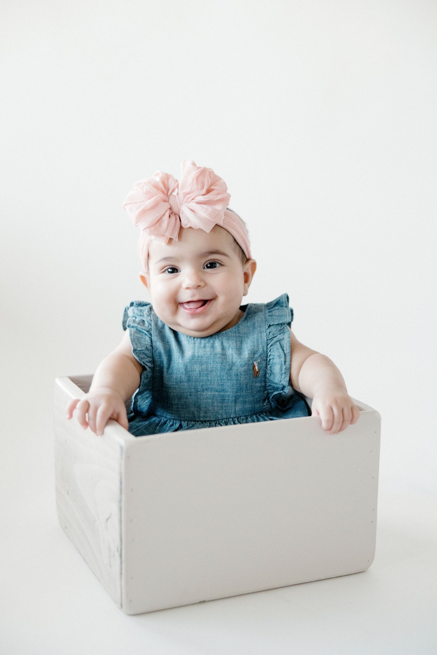 Sweet baby wearing denim dress and pink bow sits in white box during portrait session.