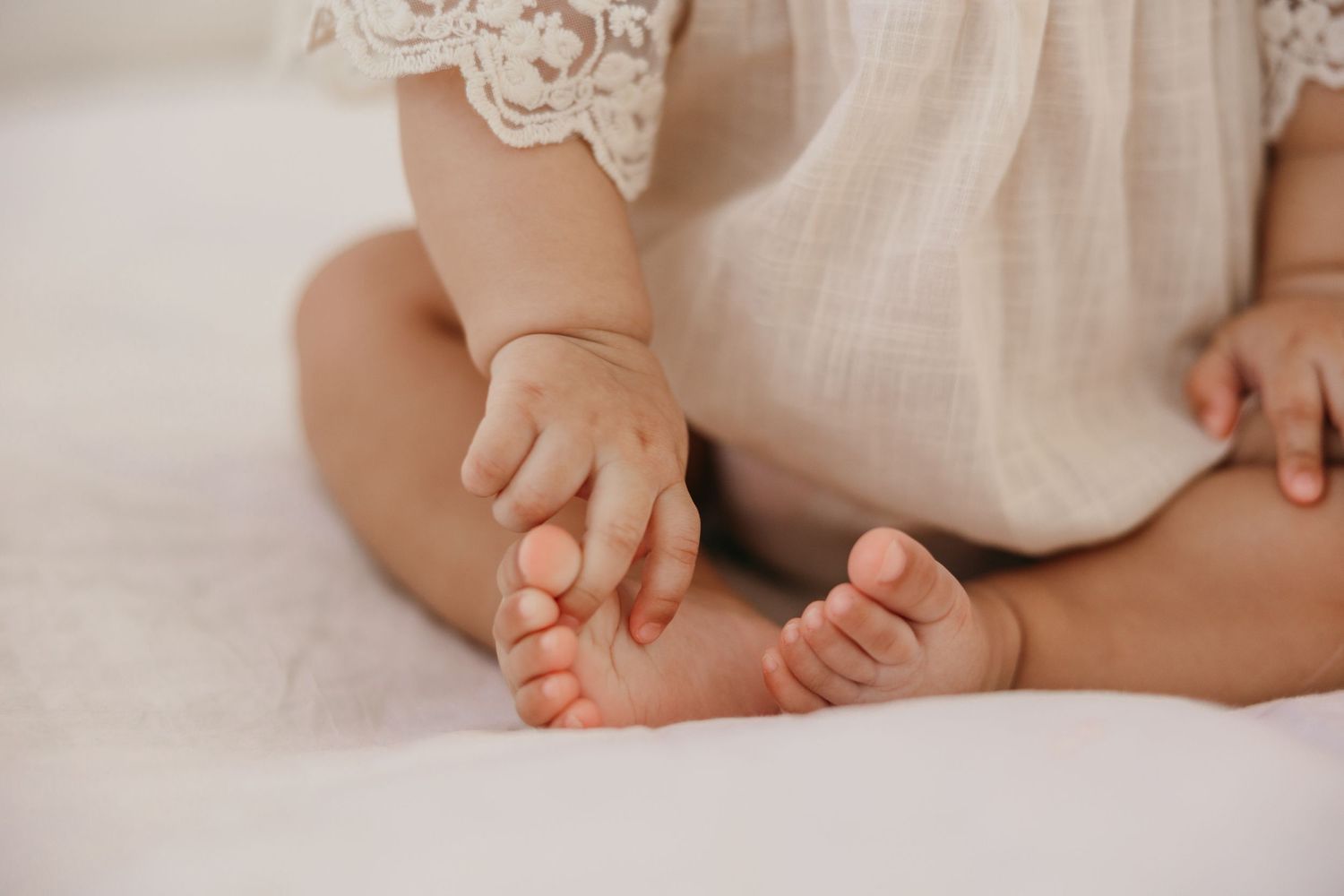 Small baby's bare feet and hands on white bedding with lace detail visible in soft natural light.