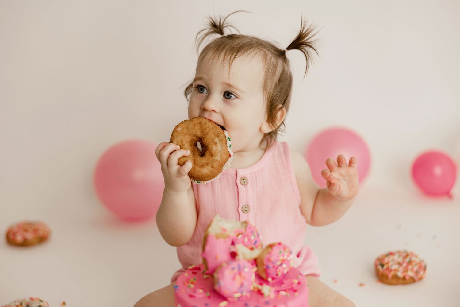 A toddler in pink enjoys donuts during a photo session with pink balloons and frosted treats in the background.