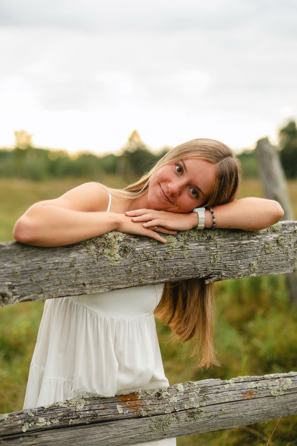 A dreamy outdoor portrait shows someone in a white dress resting on a rustic wooden fence at sunset.