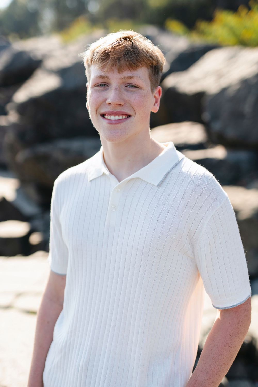 A person in a white collared shirt smiles while standing outdoors against a rocky coastal background.