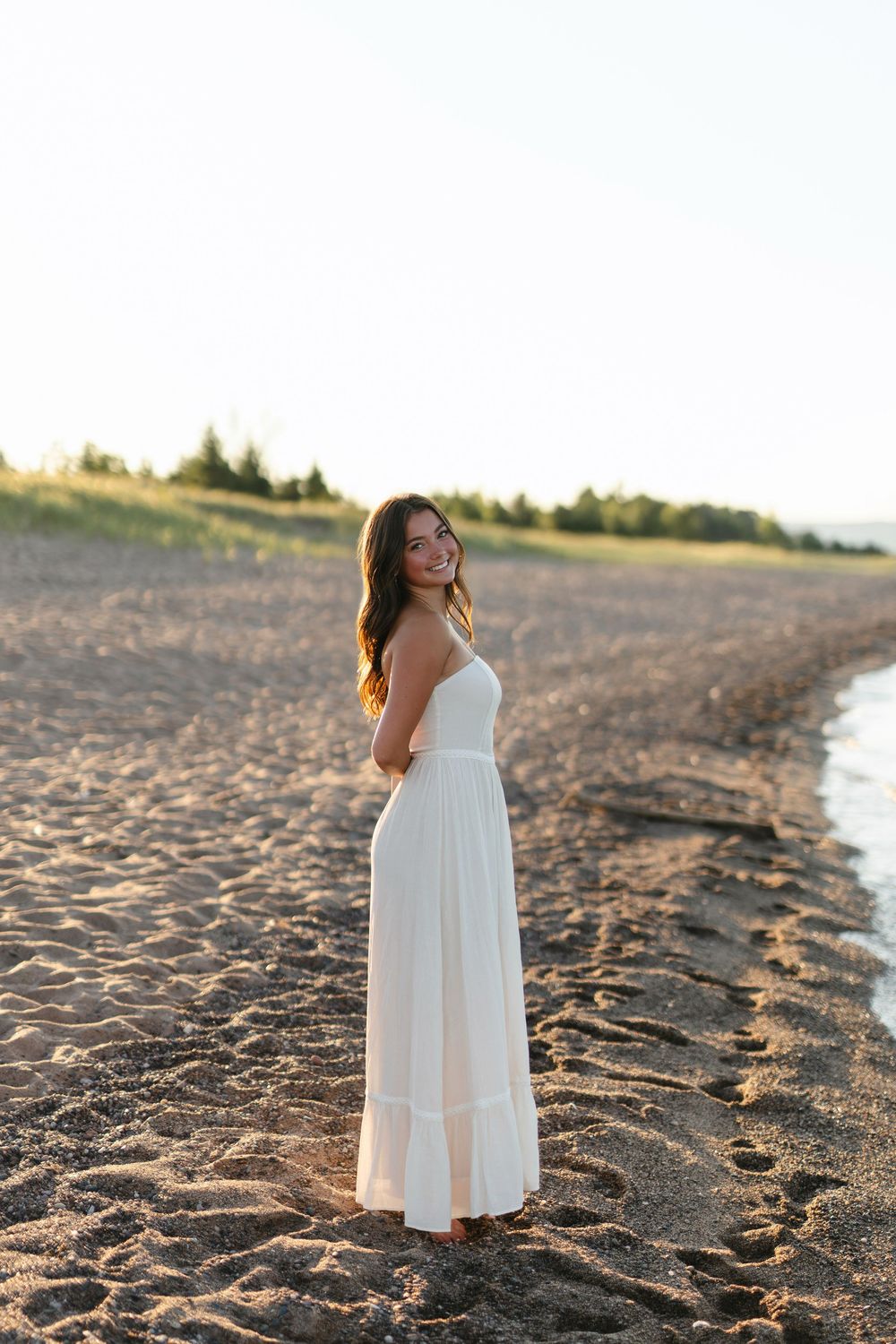A woman in a flowing white maxi dress stands on a sandy beach at golden hour.