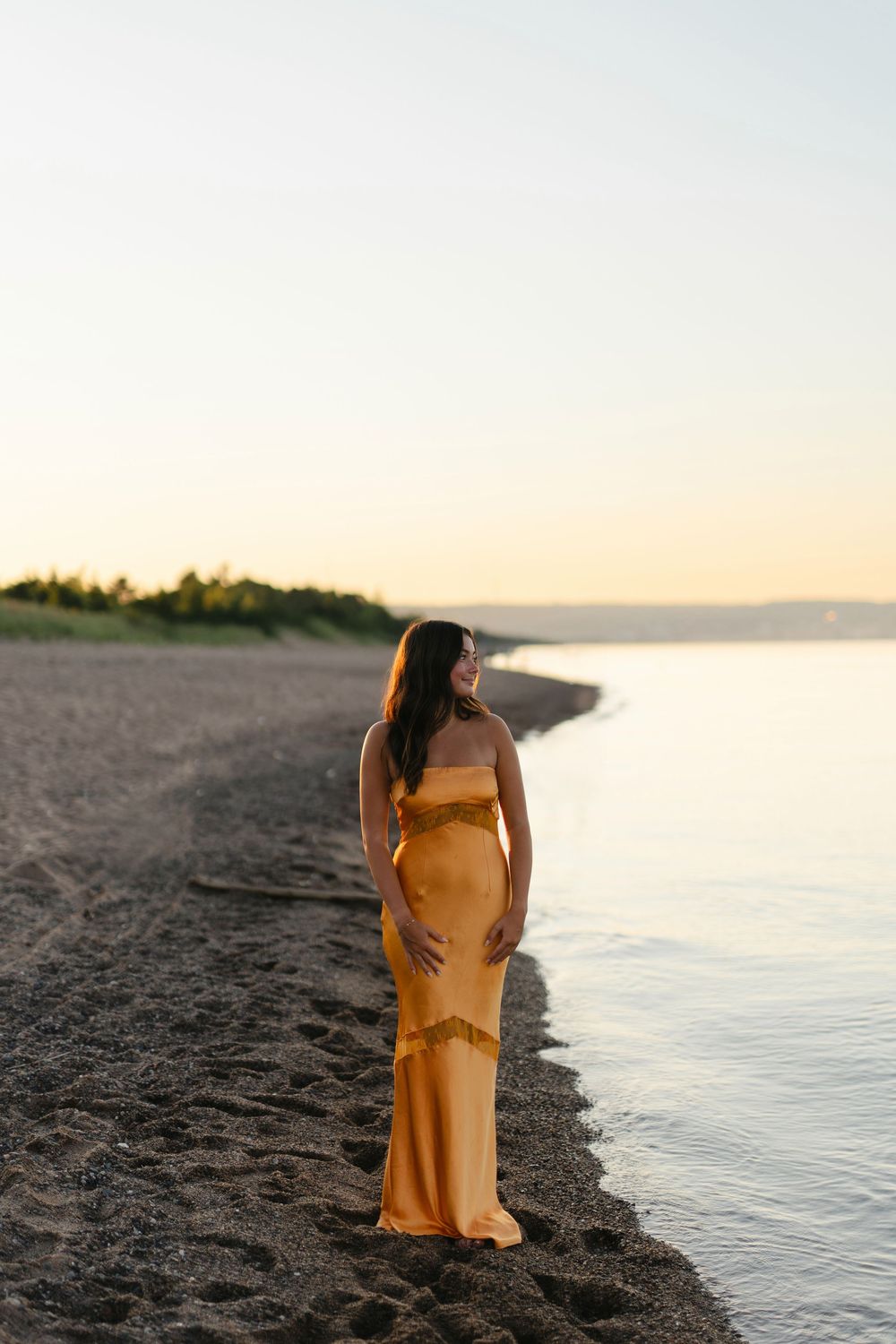 A person in a yellow dress stands on a dark sandy beach at sunset beside calm ocean waters.