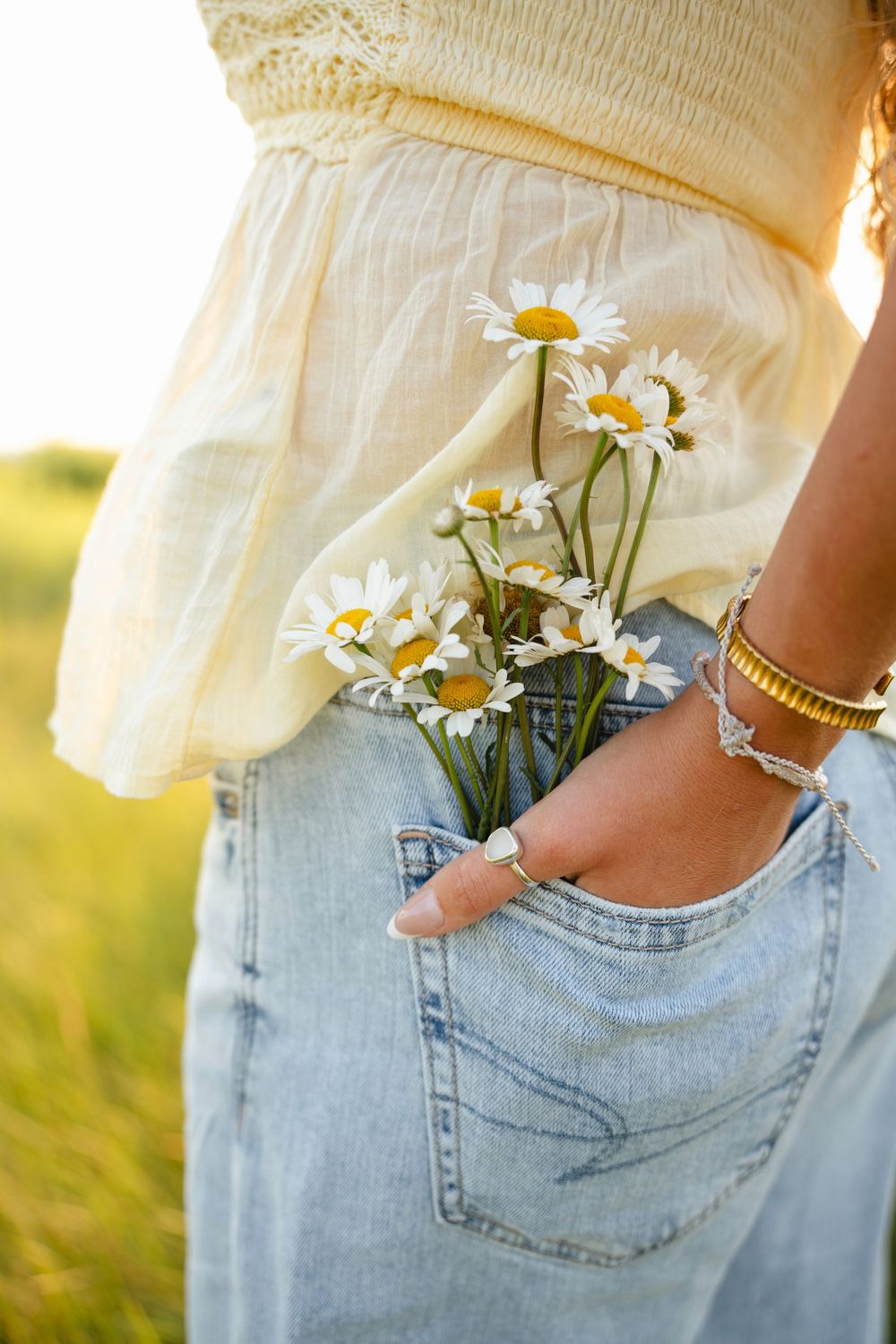 Fresh white daisies tucked into the back pocket of light wash denim jeans against a soft sunlit background.