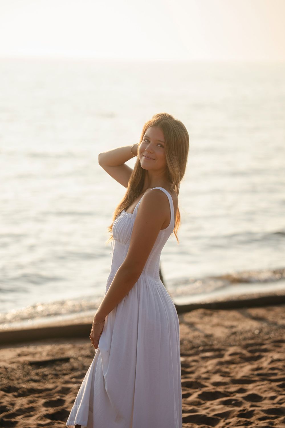 A woman in a flowing white dress poses gracefully on a sandy beach at sunset, creating a serene coastal scene.