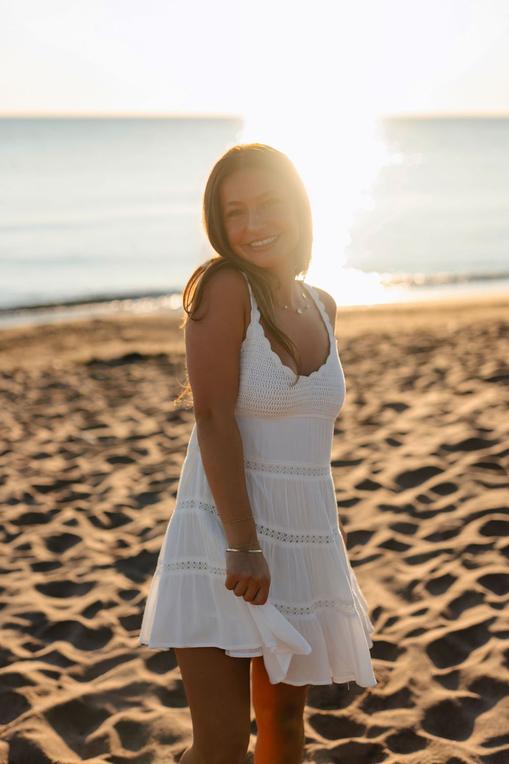 A person in a flowing white sundress walks along a sandy beach at sunset creating long shadows.