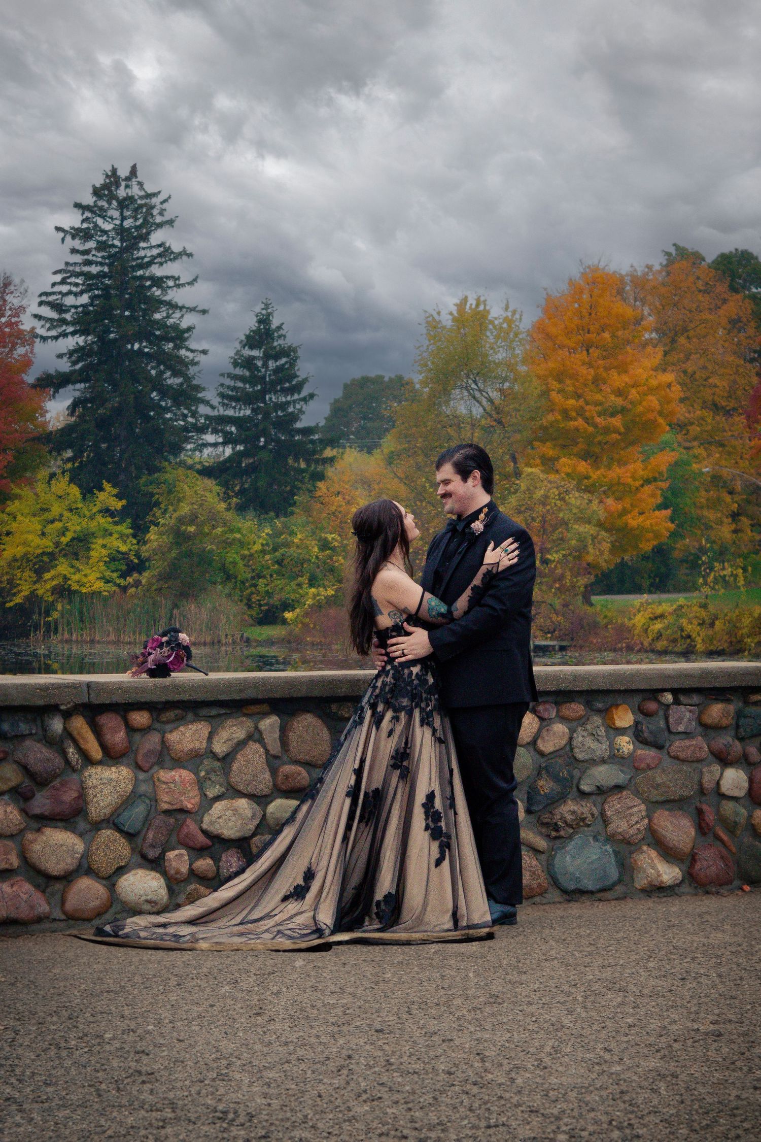 Couple in formal attire embrace on stone wall with dramatic autumn foliage background.