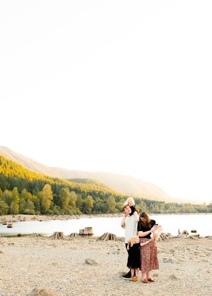 Three people embrace while standing on a rocky beach with mountains in the background.