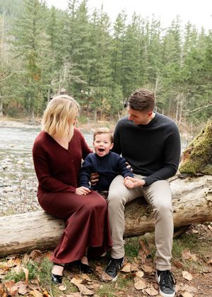 A family sits together on a fallen log by a river surrounded by pine trees.