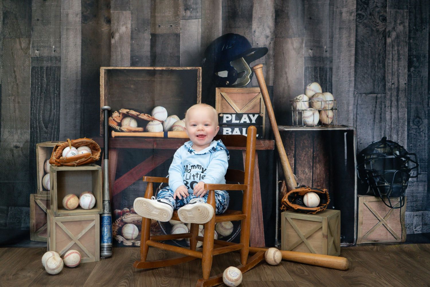 A baby sits in a wooden rocking chair surrounded by vintage wooden crates and baseball decorations.