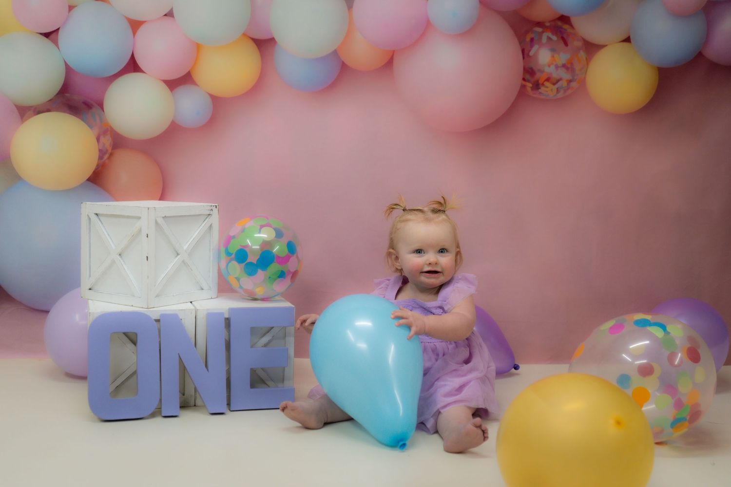 Baby in purple dress sits by colorful balloon arch and blue 'ONE' letters at first birthday photo session.