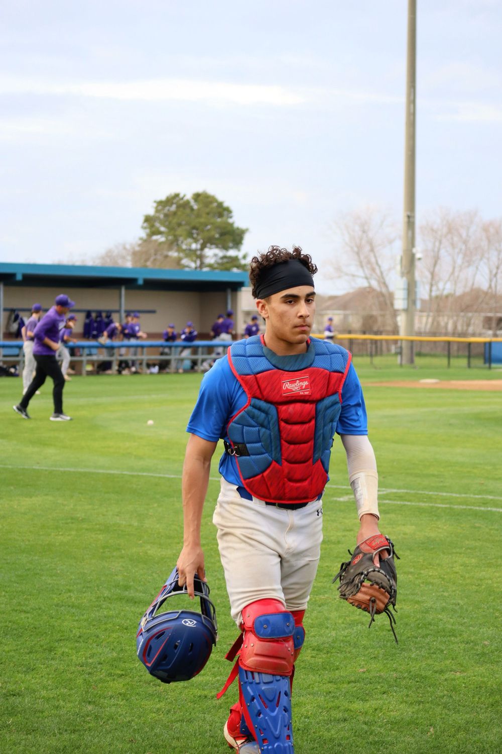 Baseball catcher in red chest protector walking across field with equipment.