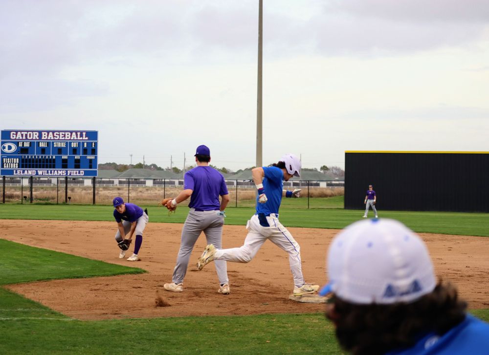 Baseball players in blue and purple uniforms practicing fielding drills on a sunny day.