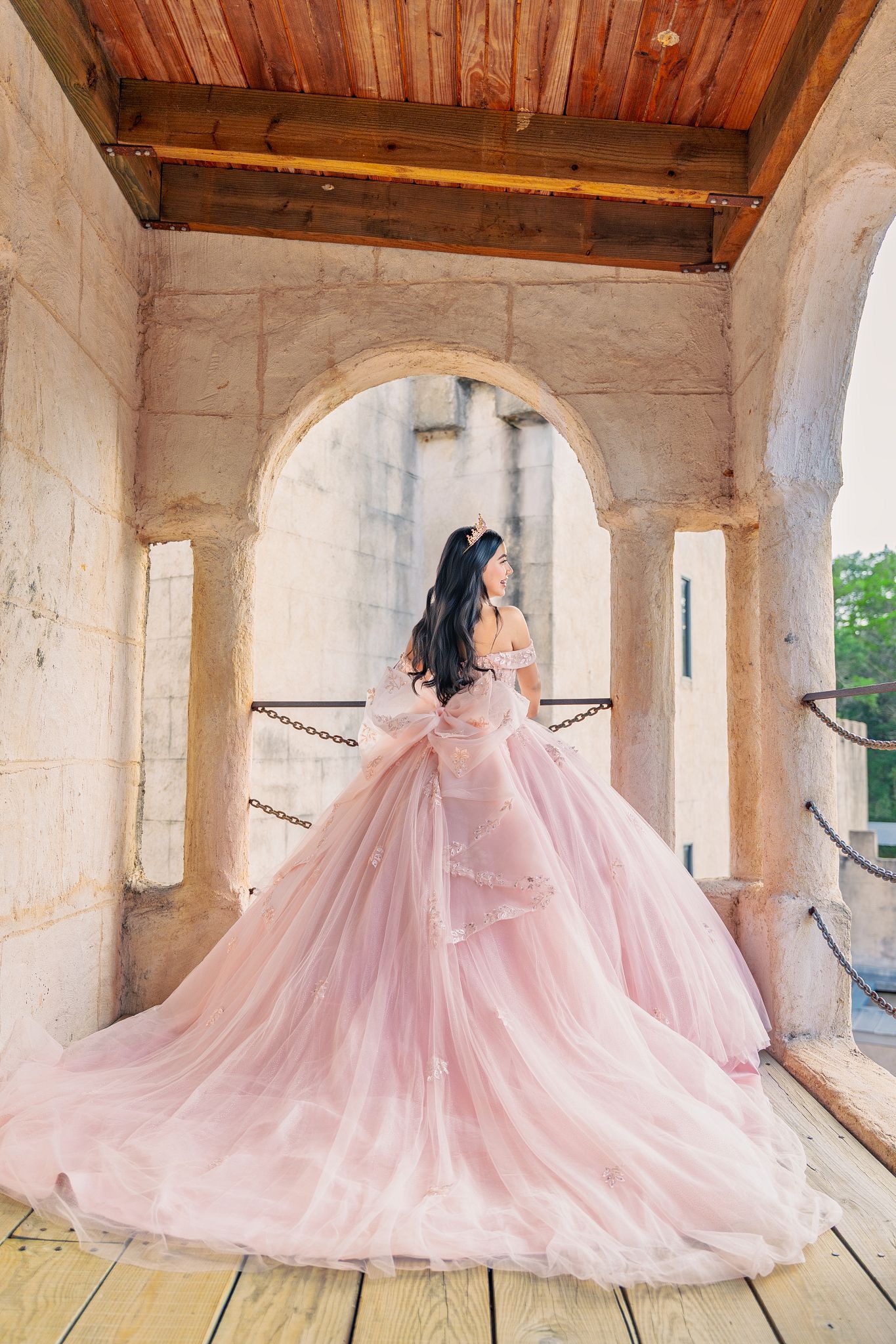 A flowing pink ball gown with tulle skirt cascades across a rustic wooden porch with stone columns.