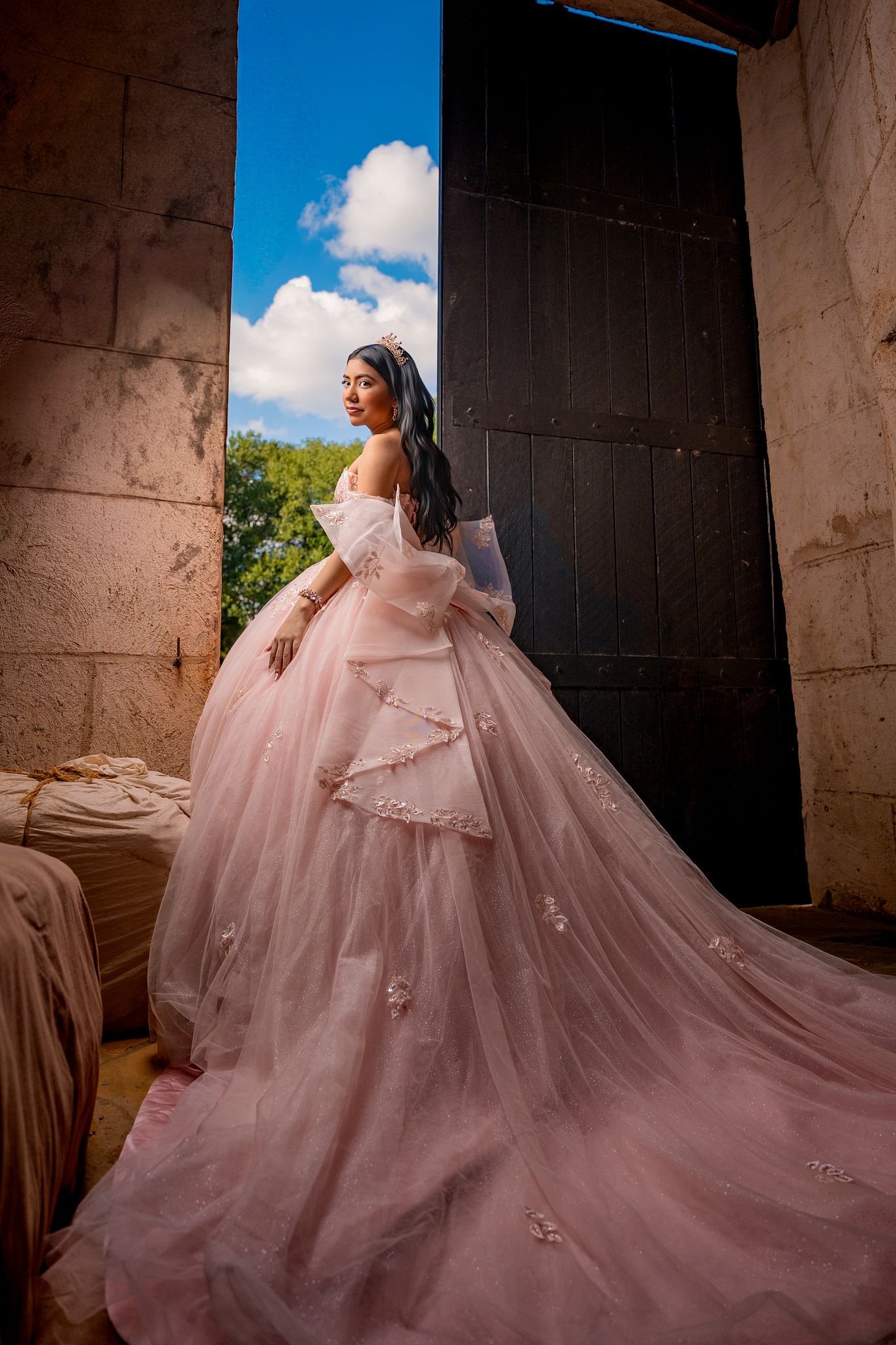 A flowing pink quinceañera gown with a dramatic train photographed against stone walls and blue sky.