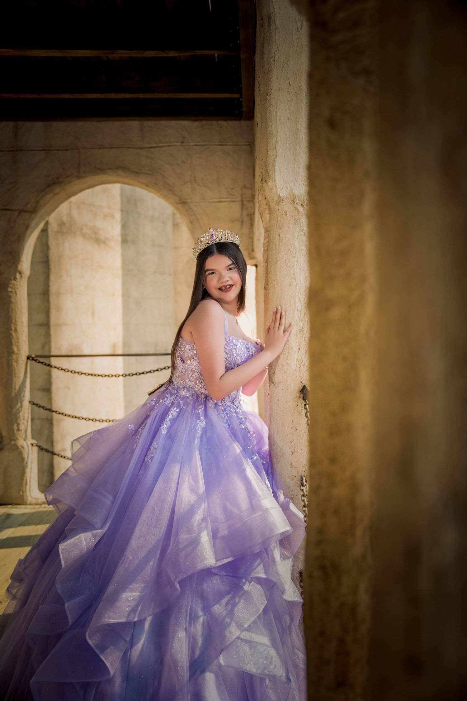 An elegant lavender quinceañera gown with floral details and ruffled layers photographed in a rustic stone archway setting.