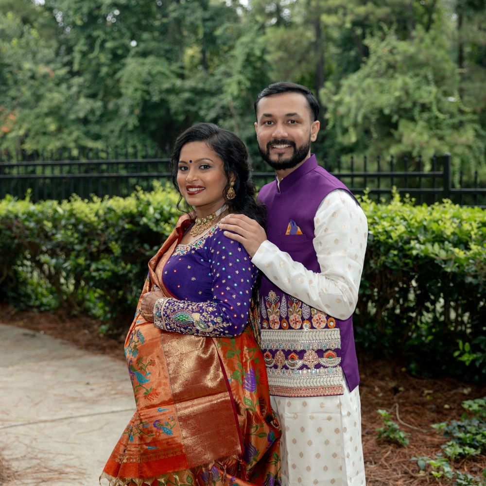 A couple in traditional Indian formal wear smiles while posing in a garden with iron fencing.