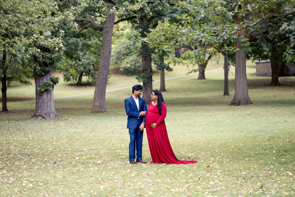 A couple poses in a scenic park setting with a striking red dress and navy suit among tall trees.