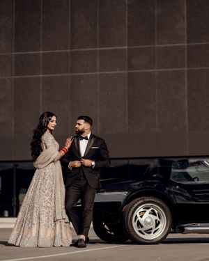 A couple in formal wear stands beside a vintage black car in an urban setting.