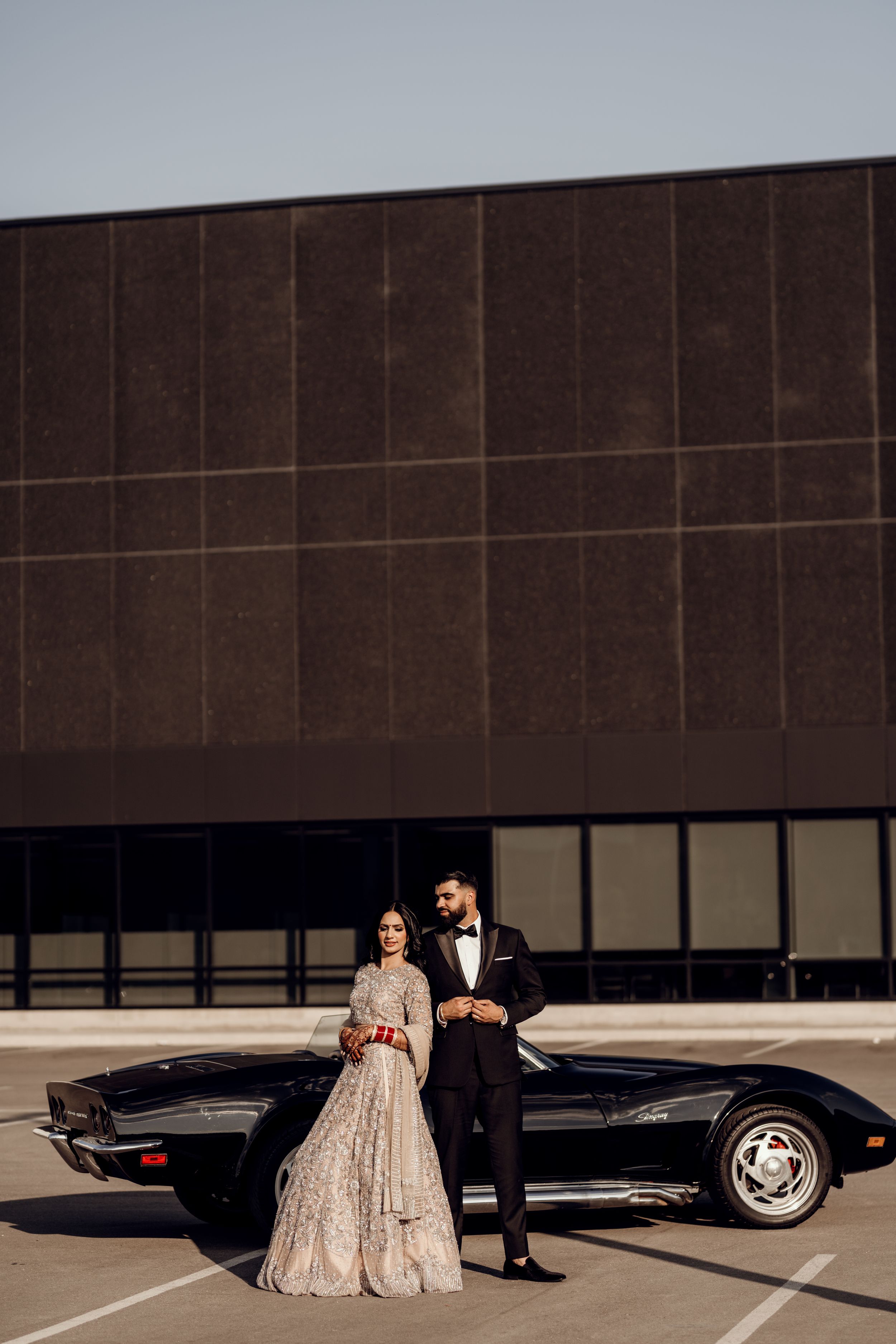 A formal couple poses with a classic black car against a modern building exterior.